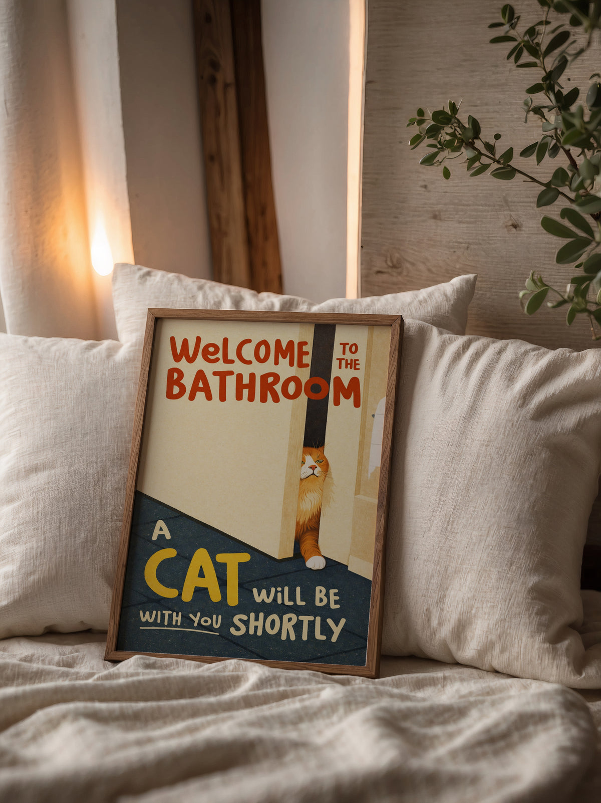 Bathroom Waiting Cat affisch i träram lutad mot kuddar i mysig sovrumsmiljö – Bozetto inredningskonst